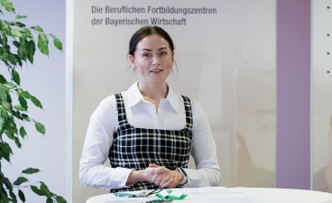persönliche Erfahrungen in der Anerkennungsberatung Auf diesem Bild ist eine junge Frau an einem Stehtisch in einem Büro zu sehen. Sie trägt ein kariertes Kleid und schaut in die Kamera. Hinter ihr ist ein Informationsplakat sichtbar, das den Schriftzug "Die Beruflichen Fortbildungszentren der Bayerischen Wirtschaft" trägt.